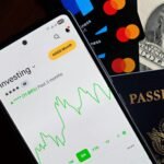 Smartphone displaying investing app, with credit cards, cash, and passport nearby, symbolizing finance